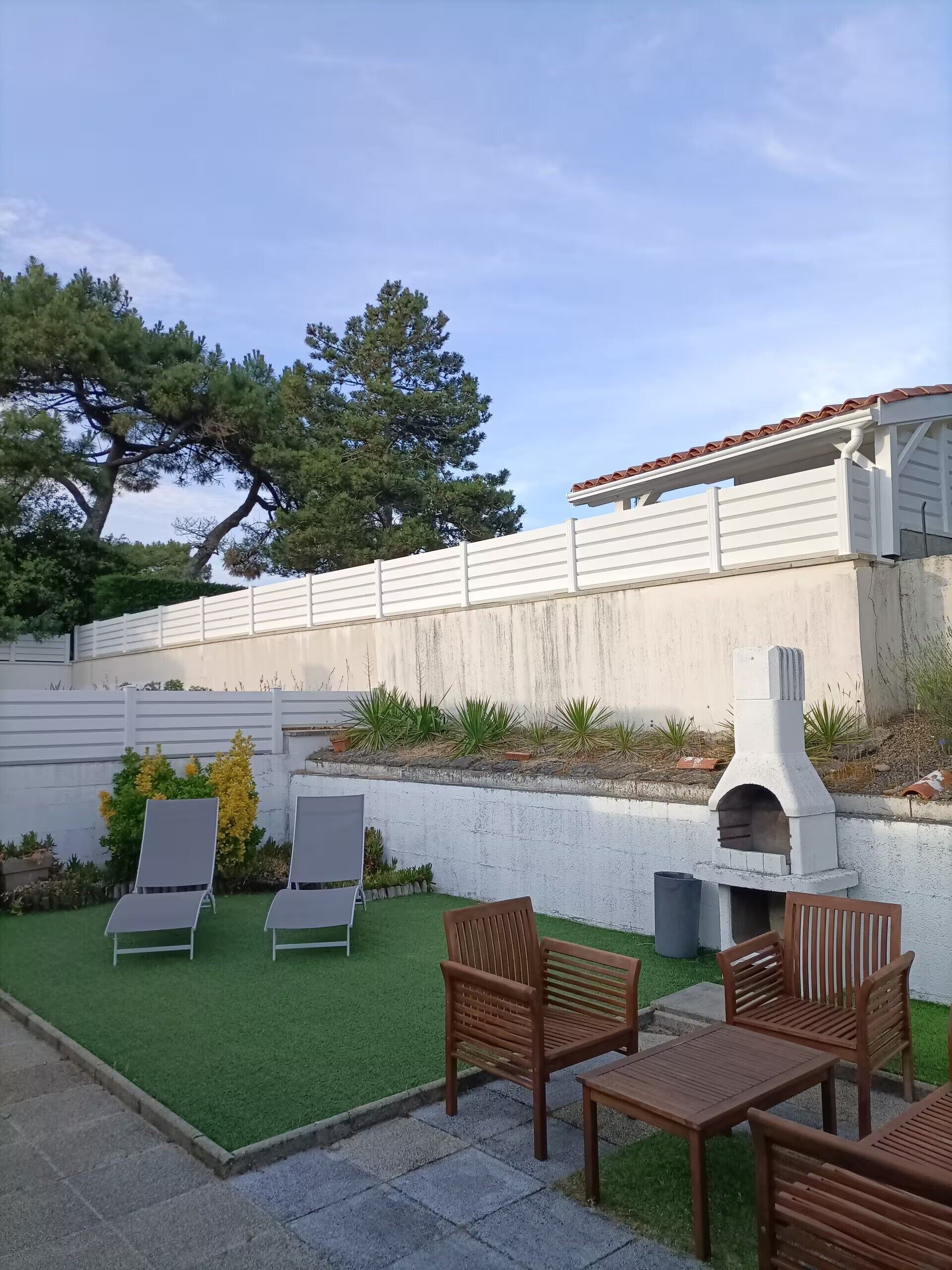 Un espace patio avec des sièges d’extérieur, un barbecue et un mur blanc surmonté d’un toit en tuiles.