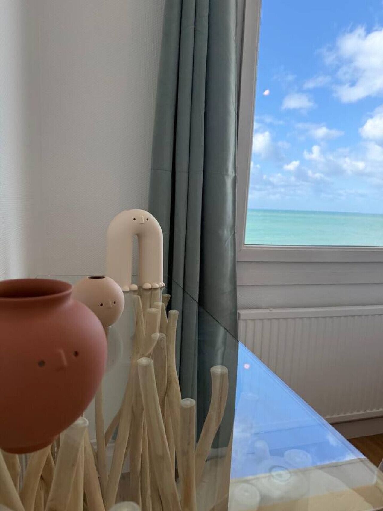 Une pièce avec vue sur l’océan, une table en verre ornée de sculptures en bois et une fenêtre avec des rideaux gris.