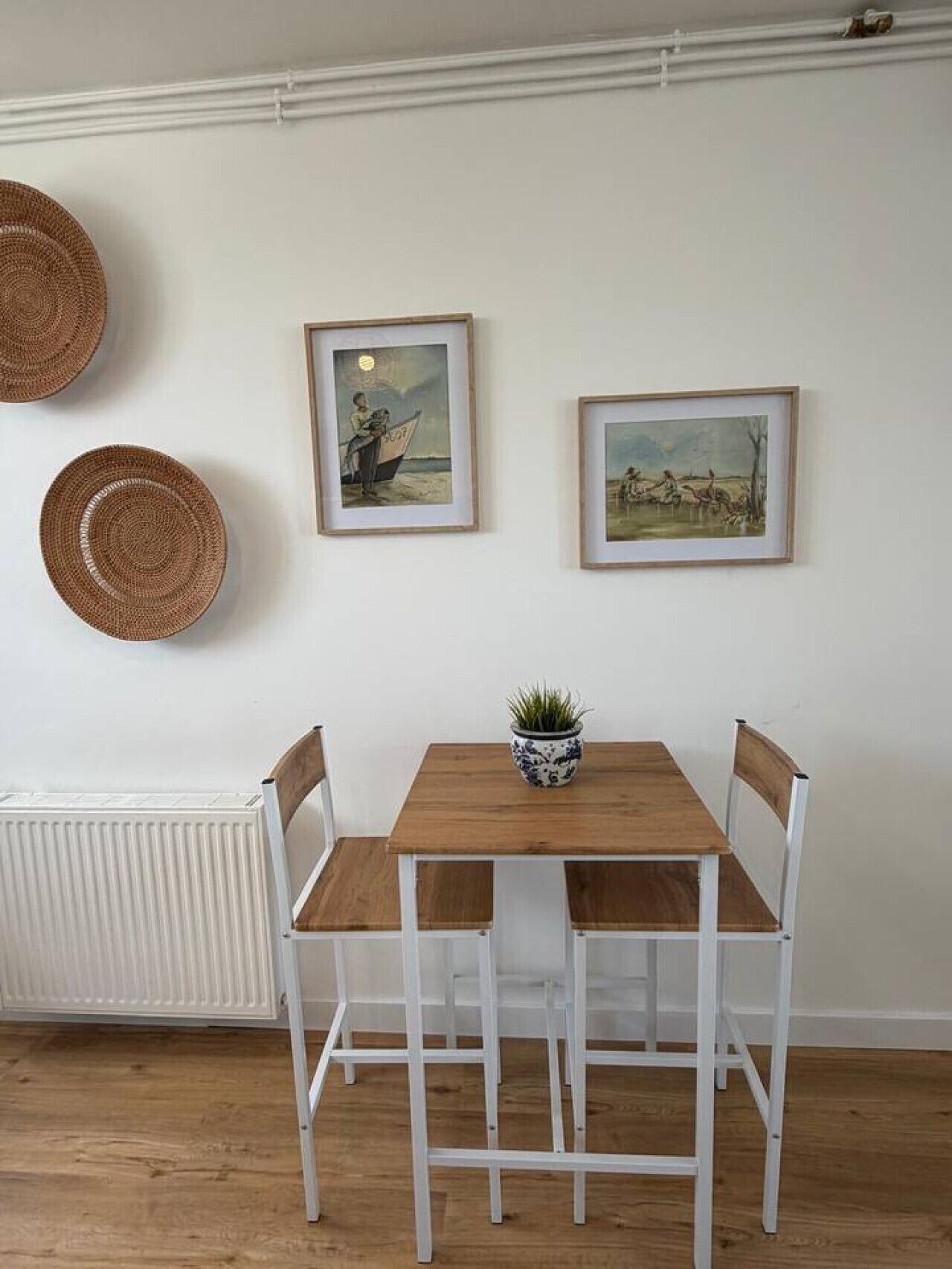 A dining area with a wooden table and white chairs, framed artwork on the wall, and woven baskets.