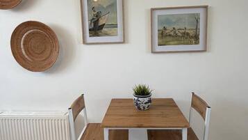 A dining area with a wooden table and white chairs, framed artwork on the wall, and woven baskets.