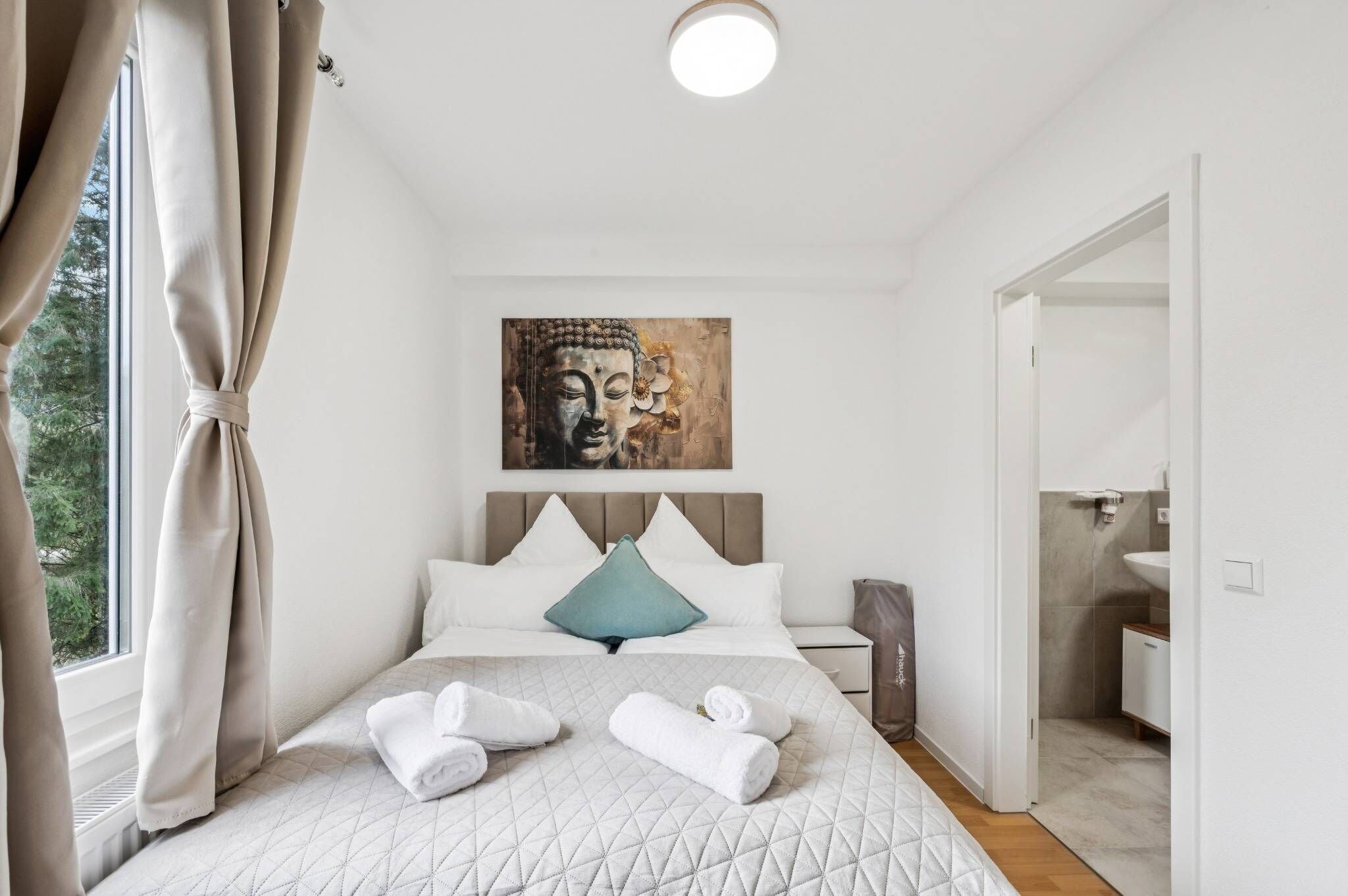 Quarto com cama, um quadro acima, janela com cortinas e banheiro visível por uma porta aberta.