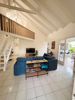 Ein geräumiges Wohnzimmer mit hoher Decke, einer Treppe, einem großen blauen Sofa, einem hölzernen Couchtisch und einem Fernseher.