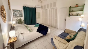 Quarto com cama, sofá, estante fixada na parede com livros, porta com venezianas e ventilador de teto.