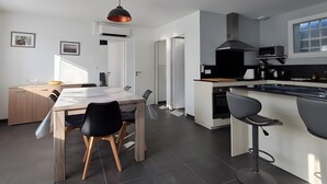 Moderni keittiö, jossa on keskeinen saareke, ruokailutila pöytineen ja tuoleineen sekä riippuvalaisin.
