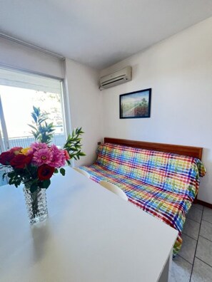 A room with a bed, a white table, a vase with flowers, and an air conditioning unit. - Flat Near the Beach With Terraces (San Michele al Tagliamento)