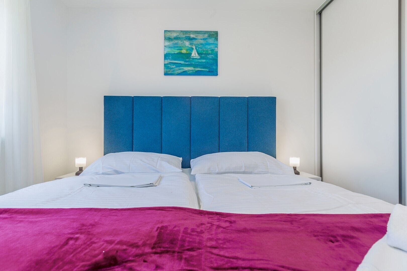 A double bed with a blue headboard, white bedding, and a pink throw blanket.