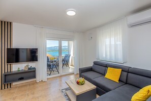 Ein modernes Wohnzimmer mit Flachbildfernseher, grauem Sofa mit gelben Kissen, Esstisch mit Stühlen und Meerblick durch große Fenster.