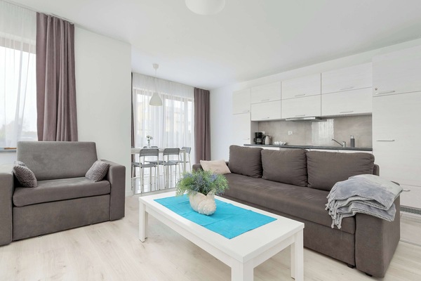 A bright and airy living area with a comfortable-looking couch, a coffee table with a decorative centerpiece, and a dining set, with a large window in the background.