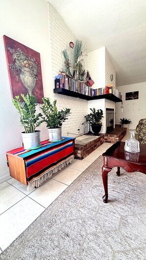 A living room with a fireplace, bookshelves, potted plants, and a large painting. - Gulf Coast Hideaway. 2BR · Near Beach & Downtown (New Port Richey)