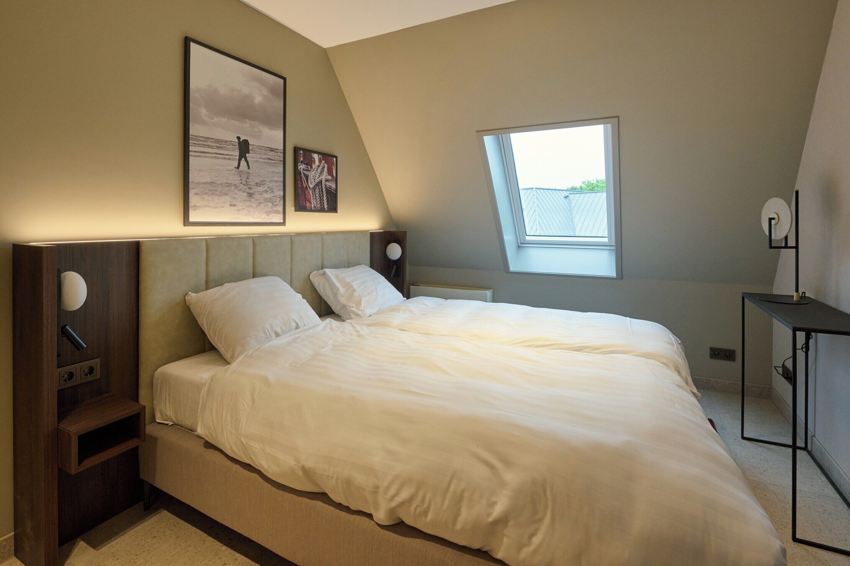 Une chambre à coucher comprenant un lit, une table de chevet, un bureau, une fenêtre et des œuvres d’art accrochées au mur.