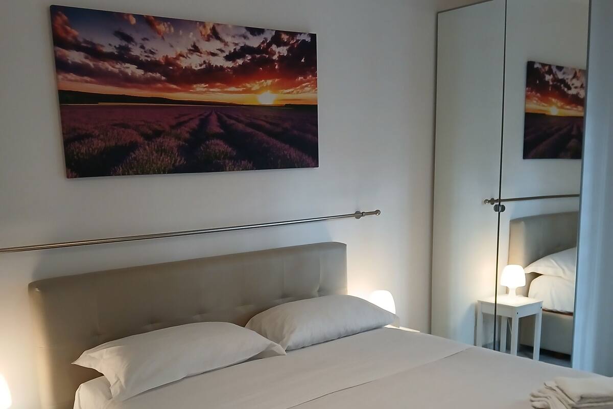 Een net opgemaakt bed met witte lakens, een nachtkastje met een lamp, een grote spiegel en een schilderij van een zonsondergang aan de muur.