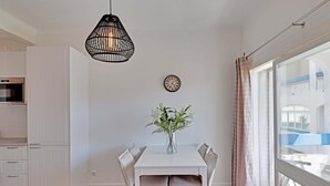 Una cocina moderna con armarios blancos, una mesa de comedor con sillas, una lámpara colgante, un reloj de pared y un amplio ventanal con cortinas.