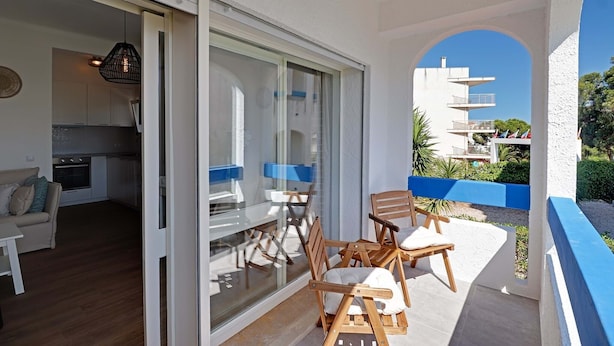 Un balcón con sillas y una mesa de madera, una zona de cocina con armarios y estufa, y una vista despejada de los edificios circundantes.