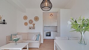 Una sala moderna con un sofá blanco, una mesita de centro y una mesa de comedor. Se ve una cocina al fondo con armarios blancos y un horno empotrado.