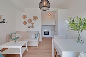 Ein modernes Wohnzimmer mit einem weißen Sofa, einem Couchtisch und einem Esstisch. Im Hintergrund ist eine Küche mit weißen Schränken und einem eingebauten Backofen zu sehen.
