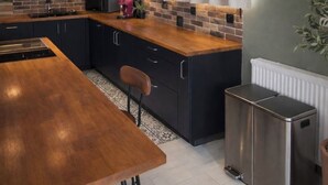 A modern kitchen with dark cabinets, a wooden countertop, a stainless steel range hood, and a built-in island with seating. - Leonard's (Bièvres)