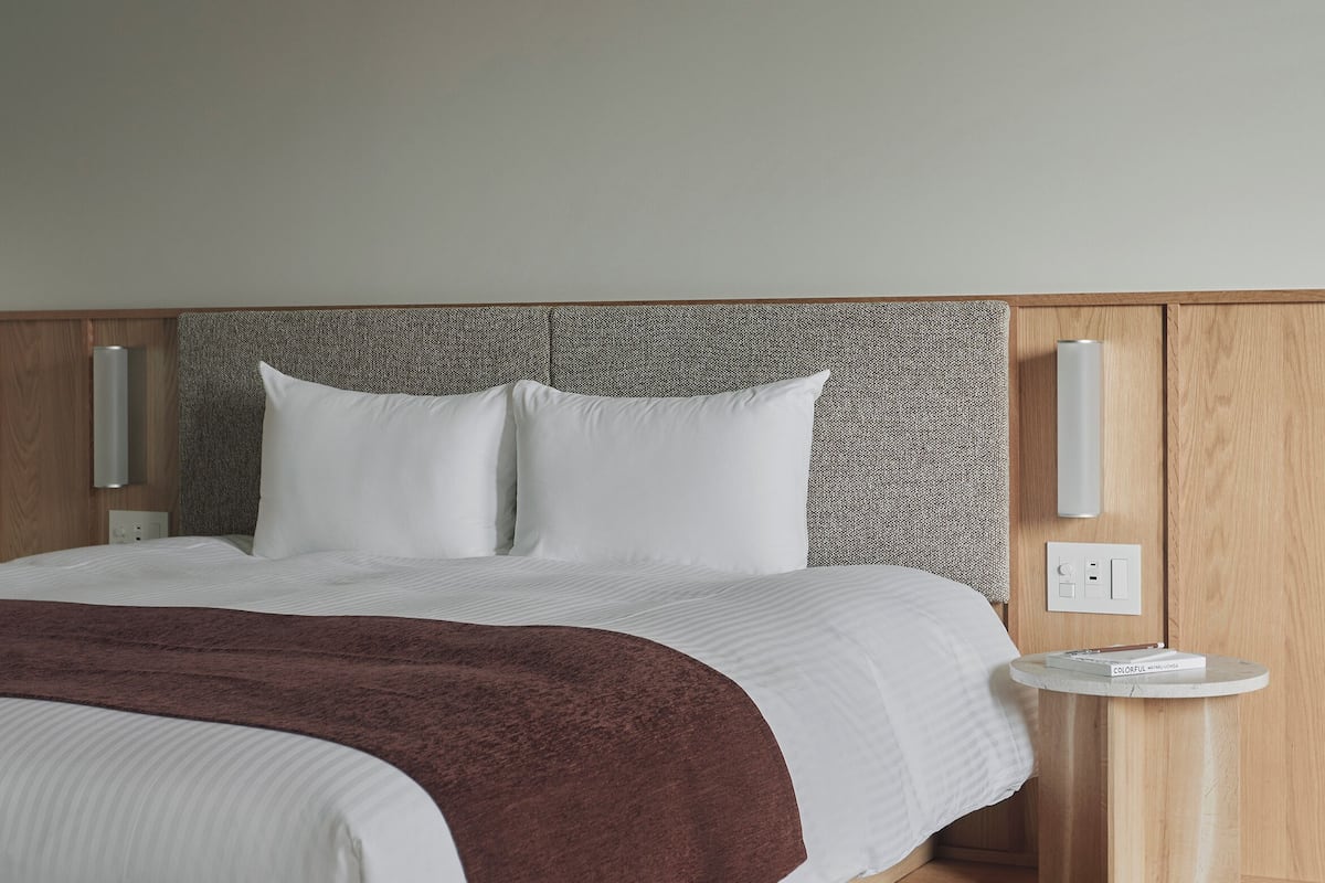 A modern hotel room with a large bed, wooden headboard, bedside tables, and a wall-mounted light.