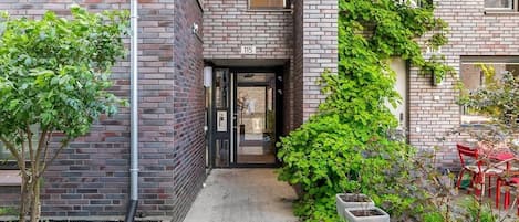A modern building with a brick facade, a covered entrance, and a small outdoor seating area with red chairs.