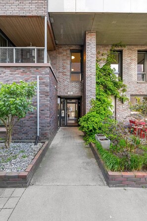 A modern building with a brick facade, a covered entrance, and a small outdoor seating area with red chairs.