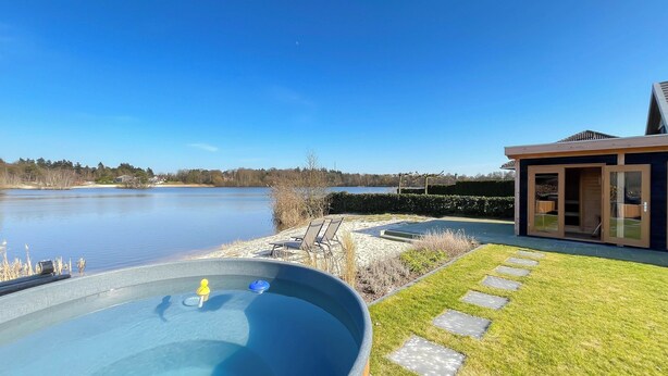 Lämmitetty poreallas, josta avautuu näkymä järvelle, nurmialue, jossa on askelmakivistä polku, ja moderni rakennus lasiovilla.
