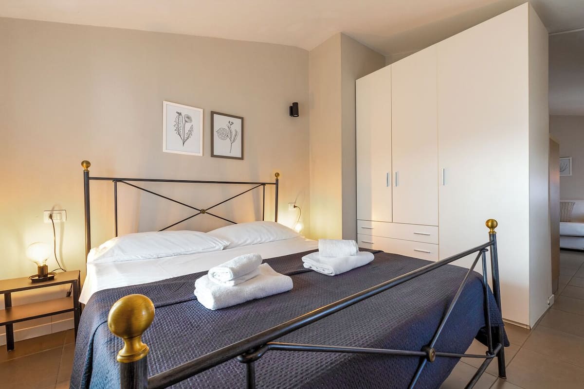 Una cama con estructura metálica, ropa de cama blanca y toallas enrolladas. Hay dos mesitas de noche con lámparas, un armario y dos ilustraciones botánicas enmarcadas en la pared.