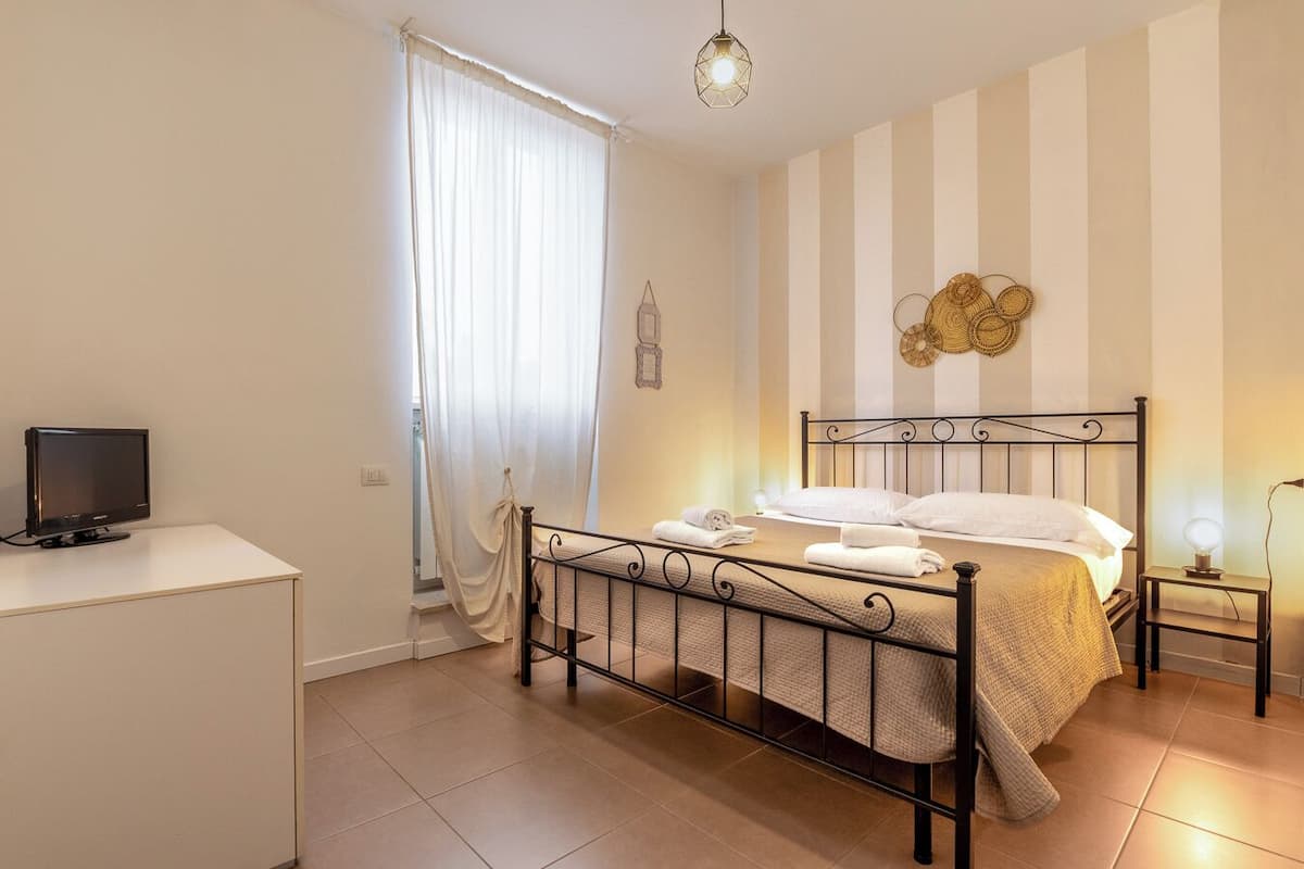 Un dormitorio con una cama, un televisor sobre un escritorio, una ventana con cortinas y una pared de papel tapiz a rayas.