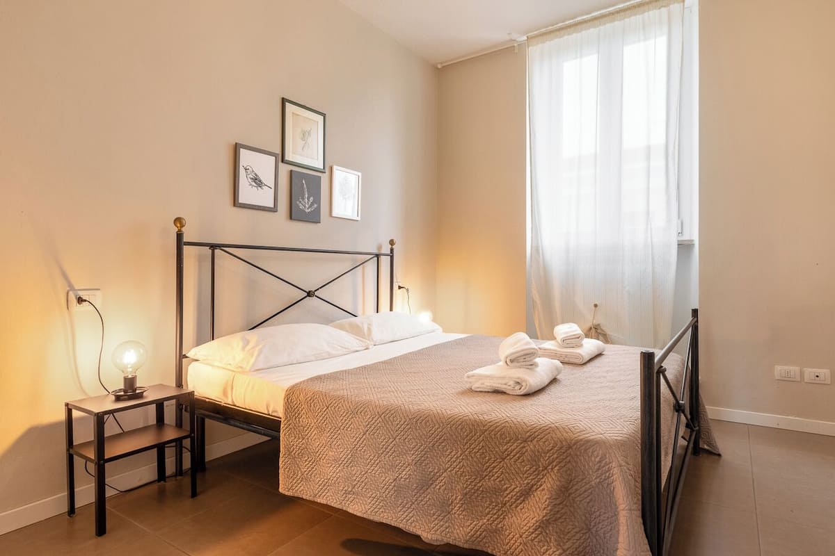 Una cama bien hecha con cabecera, una mesita de noche con lámpara y una ventana con cortinas translúcidas.