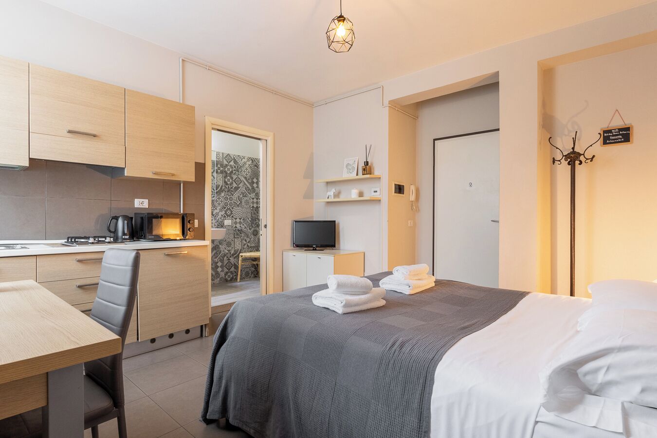 Una habitación de hotel compacta con kitchenette, una cama con ropa blanca, un escritorio y un televisor.