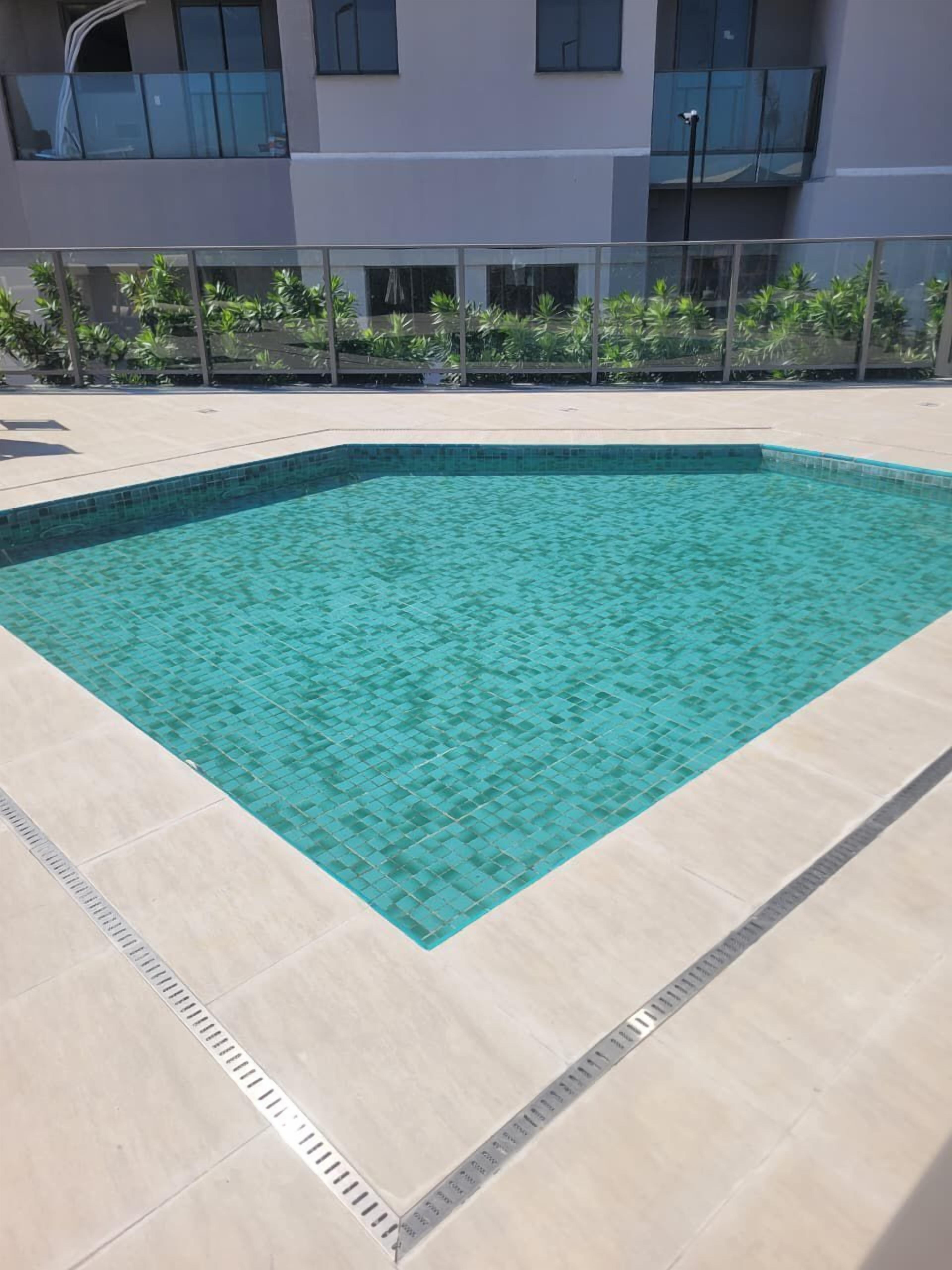Une piscine extérieure rectangulaire, dont le fond est recouvert de carreaux, entourée d’une basse clôture, avec un bâtiment aux grandes fenêtres en arrière-plan.
