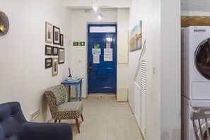 A hallway with a blue door, framed pictures, a small table with a box, and a chair. - Cosy Studio in the Heart of Valletta (Valletta)