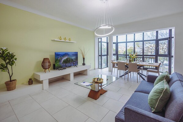 A living room with a TV, sofa, dining table, and large windows. - Spacious 3BR Retreat Overlooking Marina (Msida)