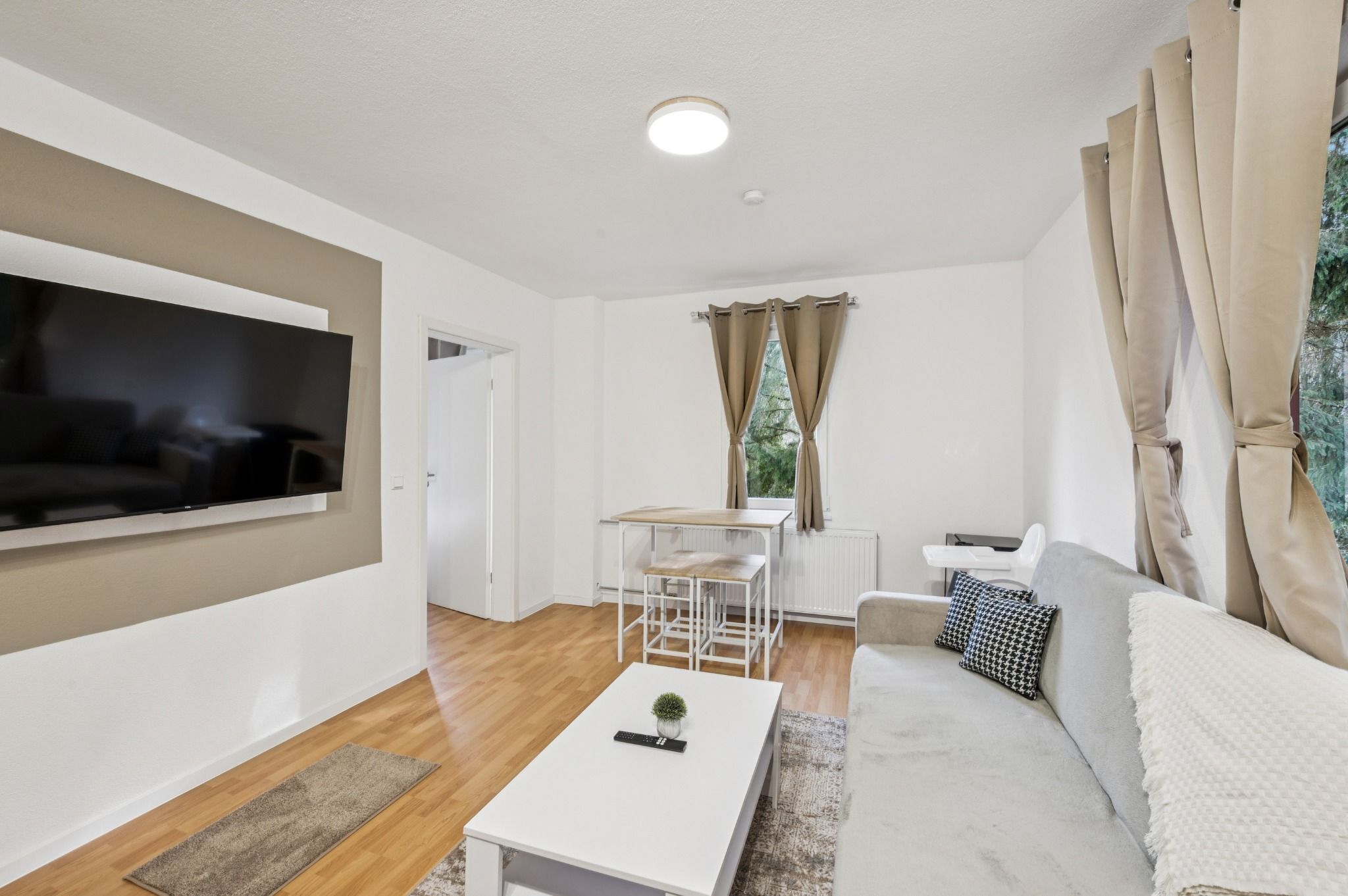 Ein modernes Wohnzimmer mit Flachbildfernseher, einer Couch, einem Couchtisch, einem Esstisch mit Stühlen und einem Fenster mit Vorhängen.
