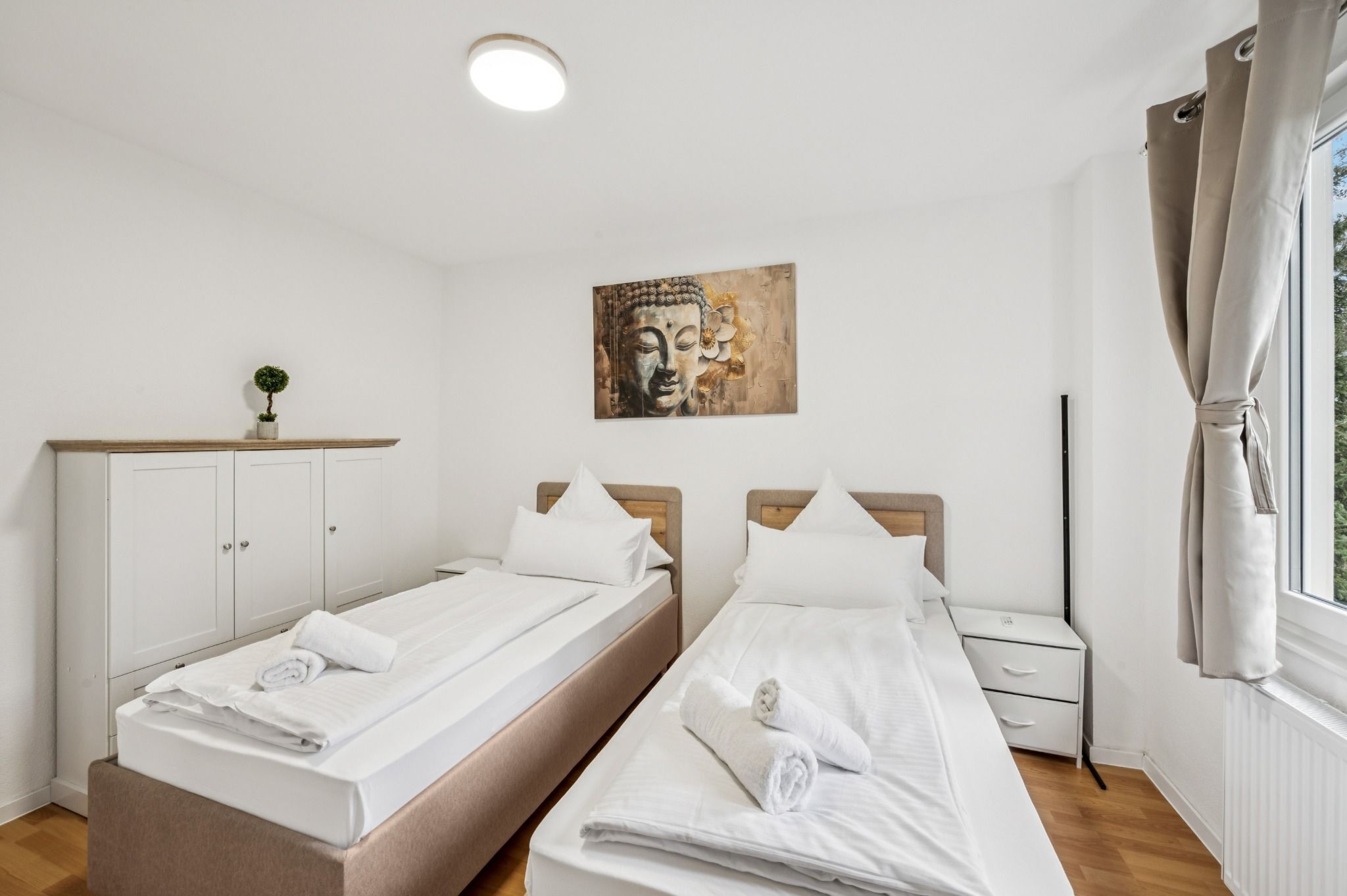 Habitación de hotel con dos camas, un armario blanco, un cuadro de Buda y una ventana con cortinas.