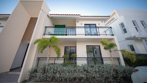 A modern two-story building with a balcony, a door, and a small garden with plants. - COZY APT 10 AIRPORT (Punta Cana)