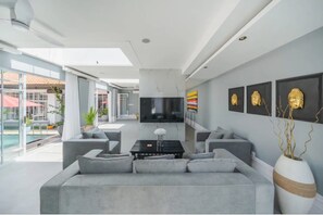Ein modernes Wohnzimmer mit einem grauen Sofa, einem Couchtisch und einem an der Wand befestigten Fernseher. An der Wand befinden sich gerahmte Kunstwerke und eine Buddha-Kopf-Skulptur.
