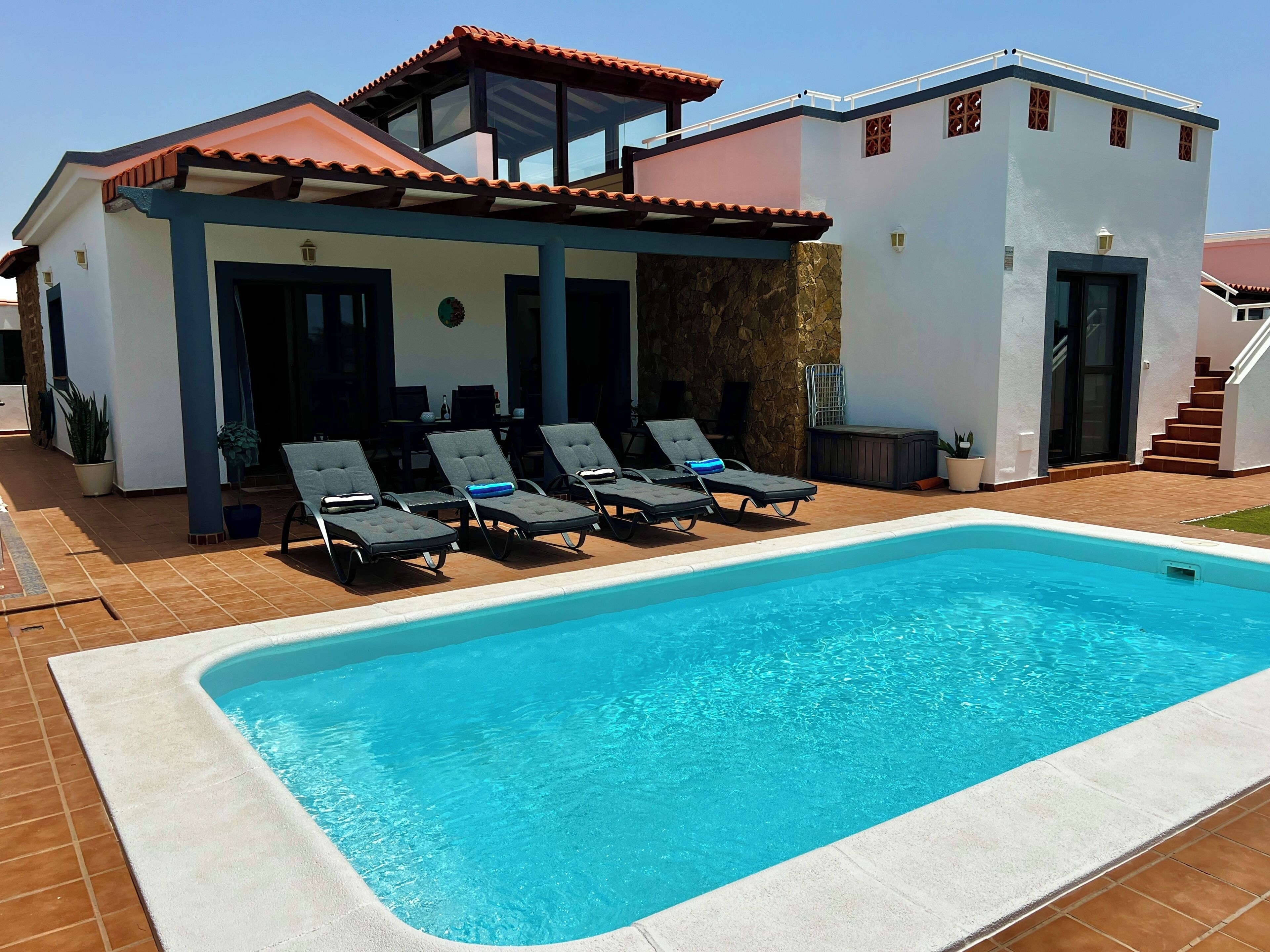 Un espace aménagé au bord de la piscine, avec des chaises longues, une piscine et un bâtiment à deux étages surmonté d’un toit en tuiles.