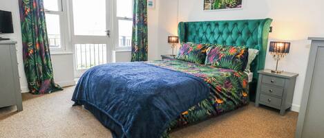 Une chambre à coucher avec une tête de lit recouverte d’un tissu vert, une couvre-lit à motifs floraux et un tableau représentant des fleurs accroché au mur.