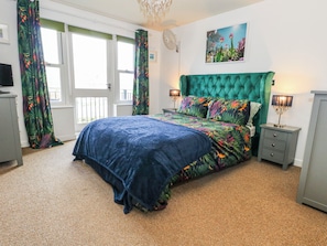 A bedroom with a green upholstered headboard, a floral bedspread, and a painting of flowers on the wall. - Rodwell Holt (Weymouth)