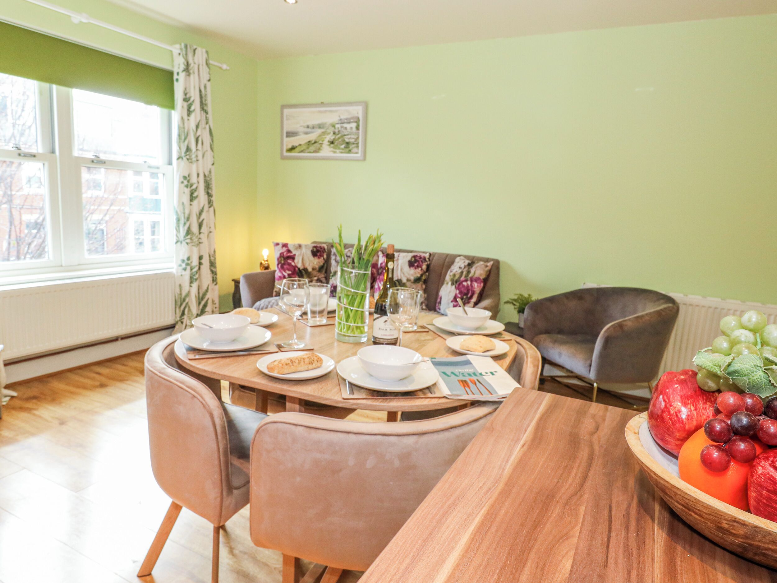 Un coin repas avec une table ronde dressée pour quatre personnes, entourée de chaises, un saladier de fruits à proximité et une fenêtre avec des rideaux.