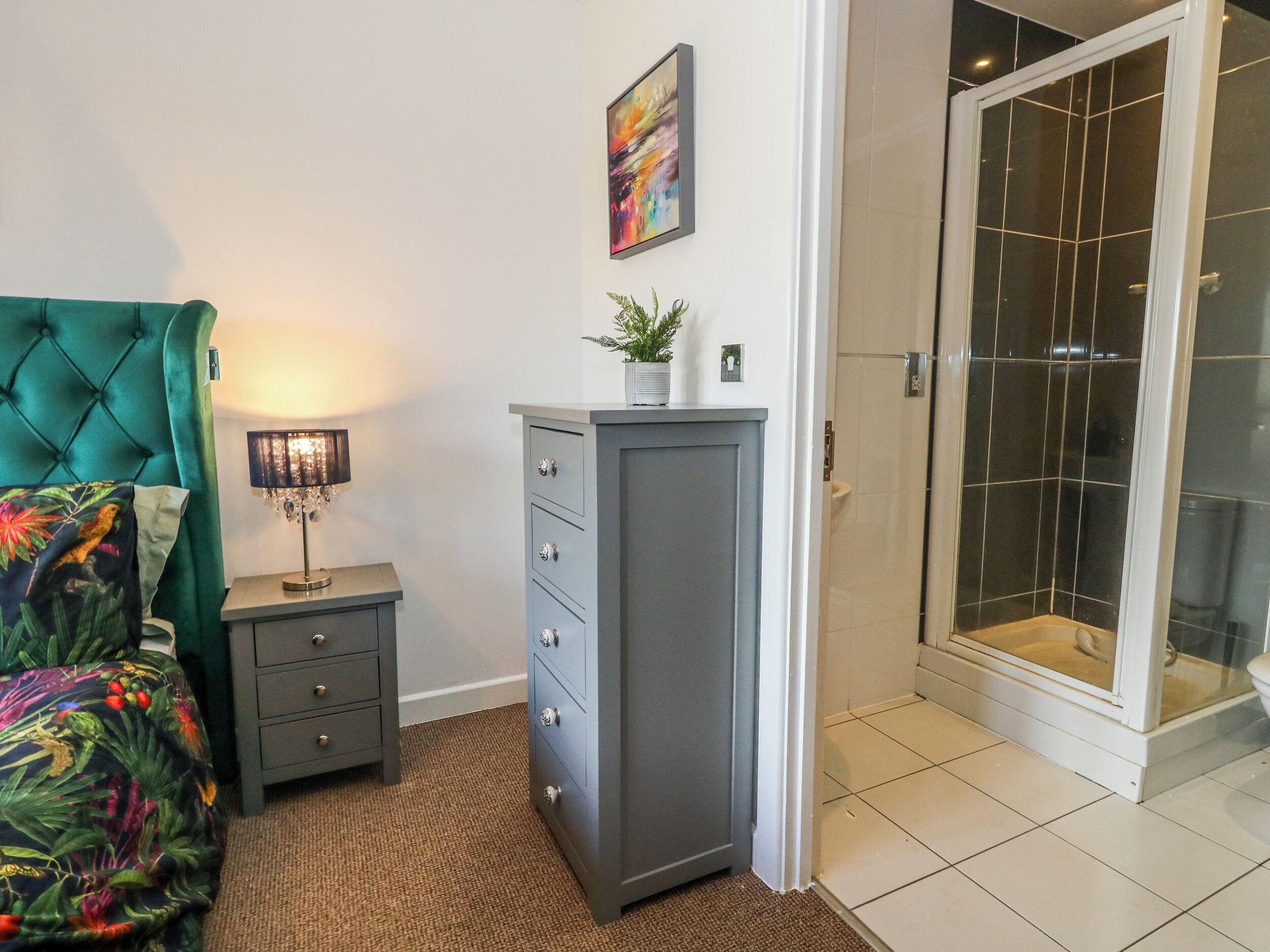 Un dormitorio con cama, mesita de noche, cómoda, lámpara, planta en maceta y ducha con puertas de vidrio.