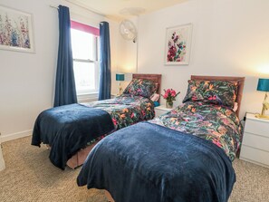 A bedroom with two beds, a window with curtains, a fan, a nightstand with a lamp, and a wall with a framed picture. - Rodwell Holt (Weymouth)