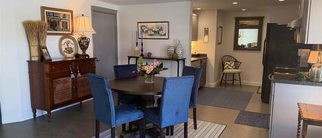 A dining area with a round table, blue upholstered chairs, a patterned rug, and a kitchen in the background.