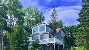 A two-story house with a balcony, surrounded by trees and a gravel driveway. - CASTLEWOOD COTTAGE - Town of Owls Head (Owls Head)