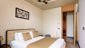 Una cama bien hecha con cabecera, una mesita de noche con lámpara, una obra de arte colgada en la pared y una puerta abierta que da a otra habitación.