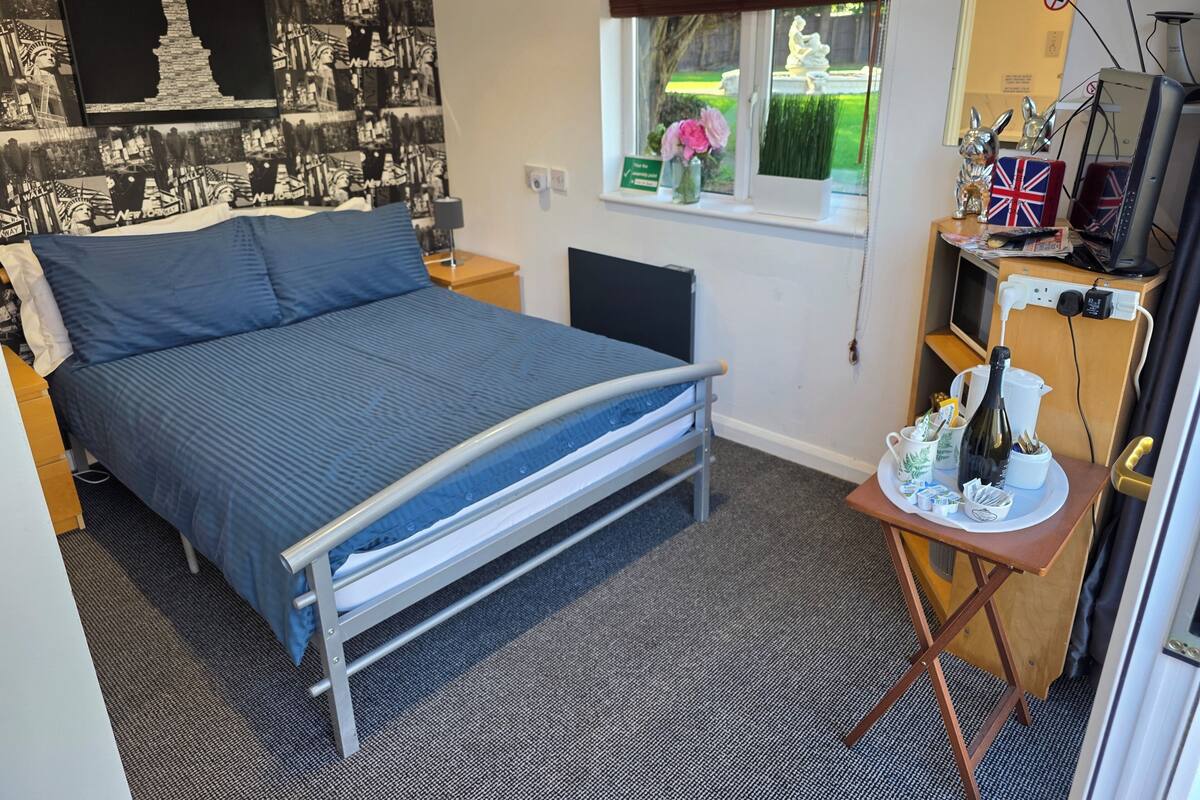 A bedroom with a bed, a desk with a computer, a window with a view of greenery, and a wall with a black and white patterned wallpaper.