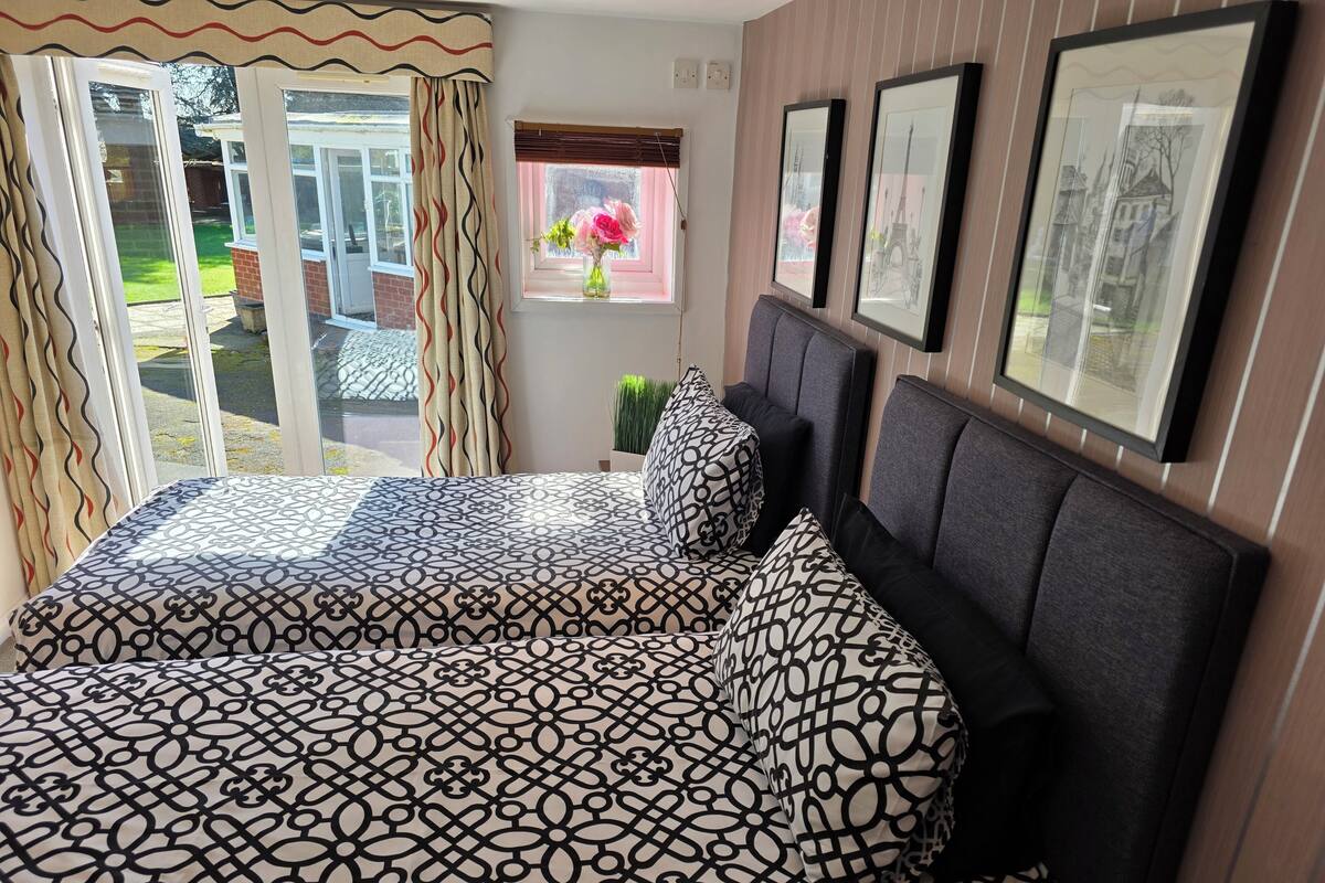 A bedroom with a large bed, a window with curtains, a small table with a vase, and framed pictures on the wall.