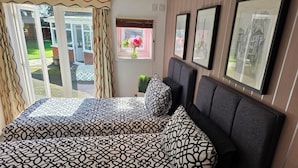 A bedroom with a large bed, a window with curtains, a small table with a vase, and framed pictures on the wall. - Redlands (Bickenhill)