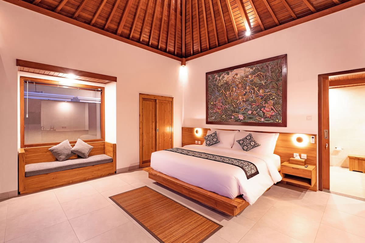 Un dormitorio con techo de madera, una cama grande con ropa blanca, un banco con cojines, una obra de arte en la pared y una mesita de noche con una lámpara.