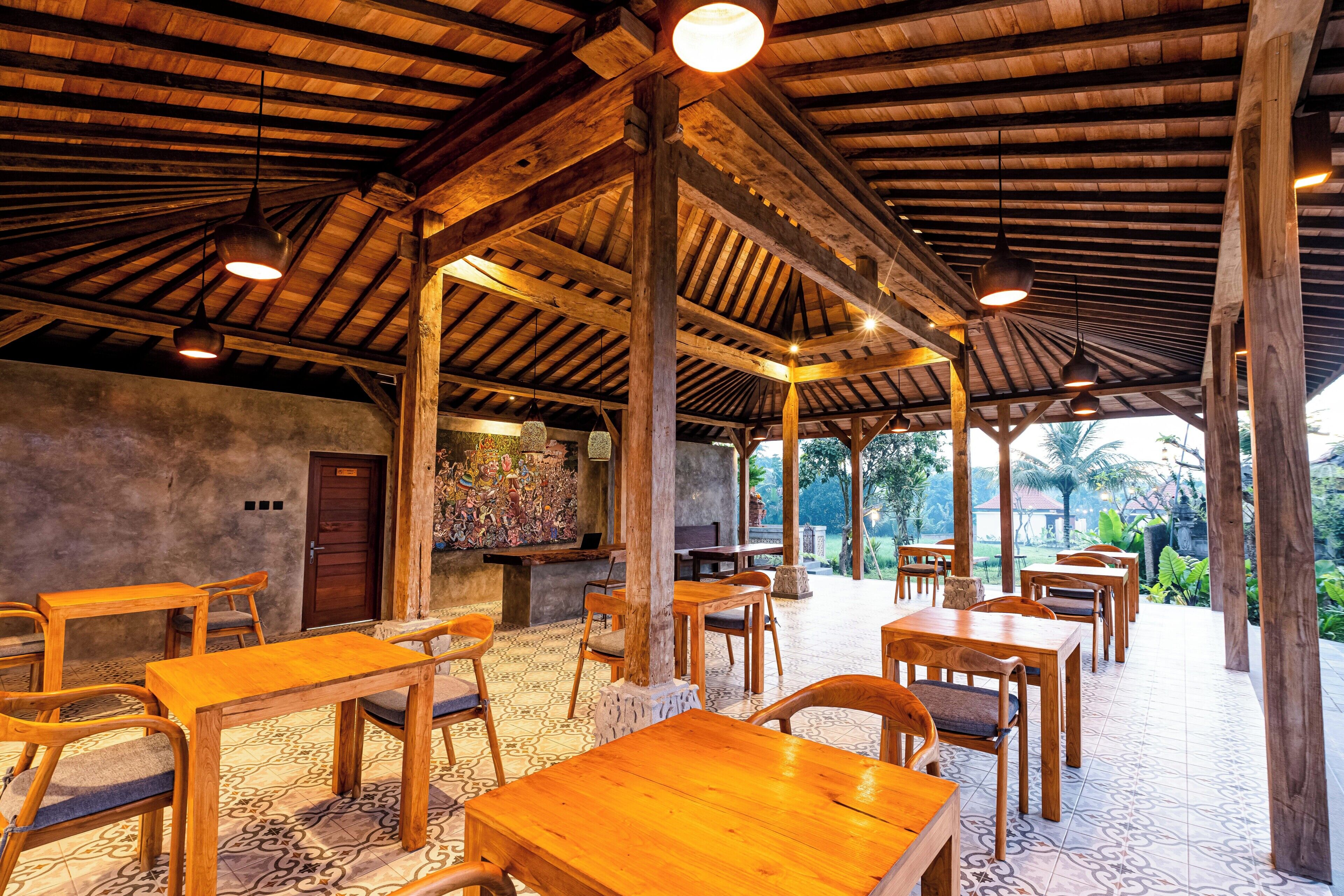 An indoor dining area with wooden tables and chairs, a high ceiling with exposed beams, and a view of greenery outside.