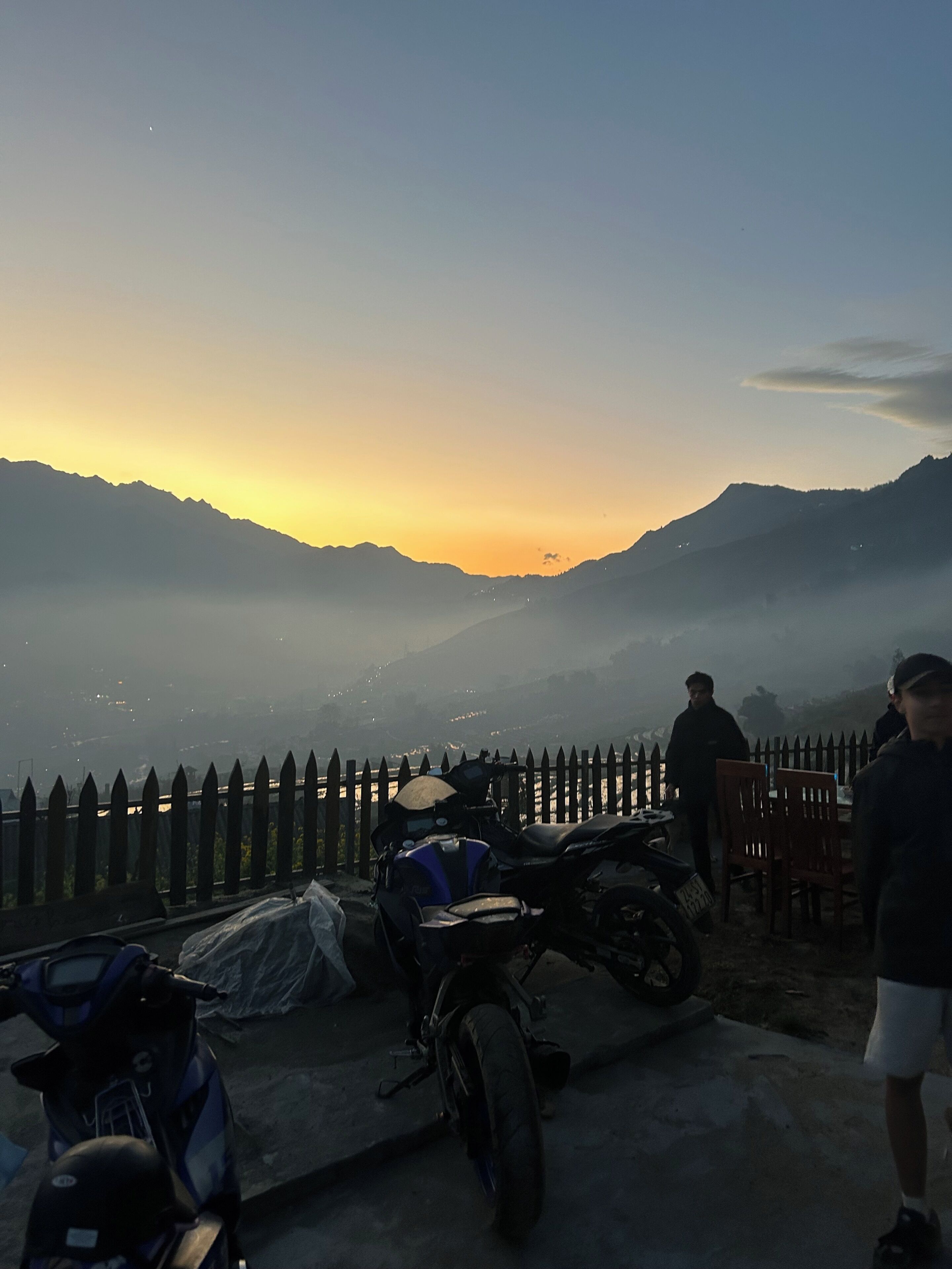 Blick auf einen Sonnenuntergang mit Bergen, einem Holzzaun und abgestellten Motorrädern.
